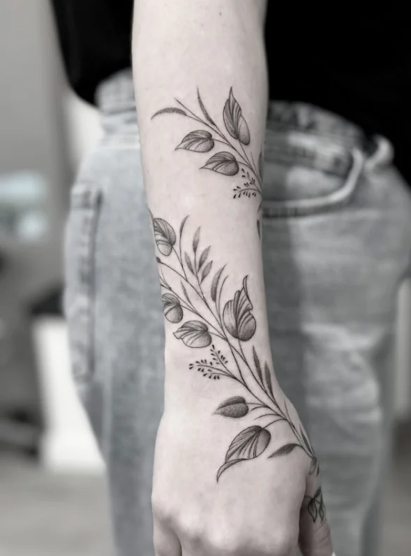 Fine-line leafy vine curving around a woman's forearm in a delicate botanical tattoo design