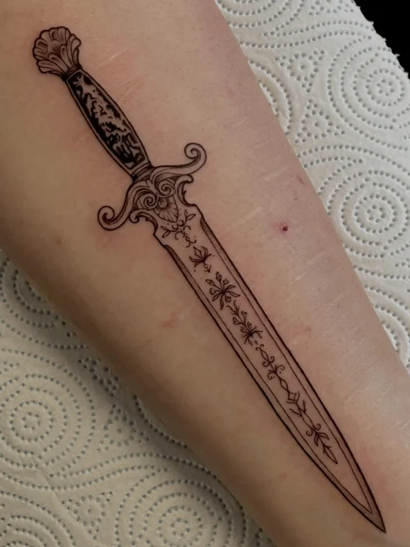 Fine-line dagger tattoo running vertically along the inner forearm