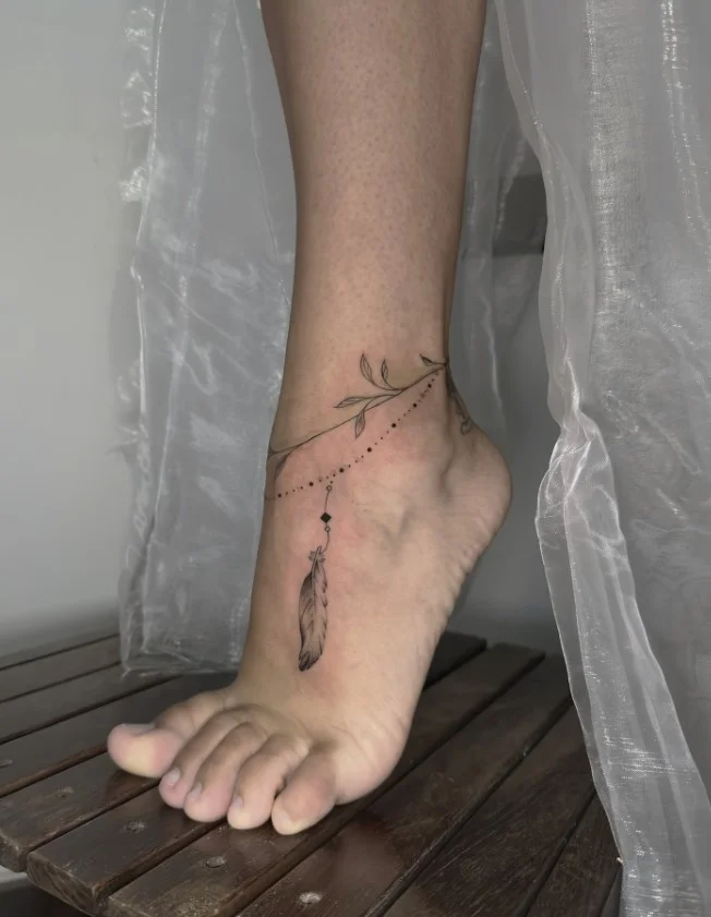 Fine line feather ankle tattoo featuring a delicate feather outline with slender details positioned vertically along the ankle