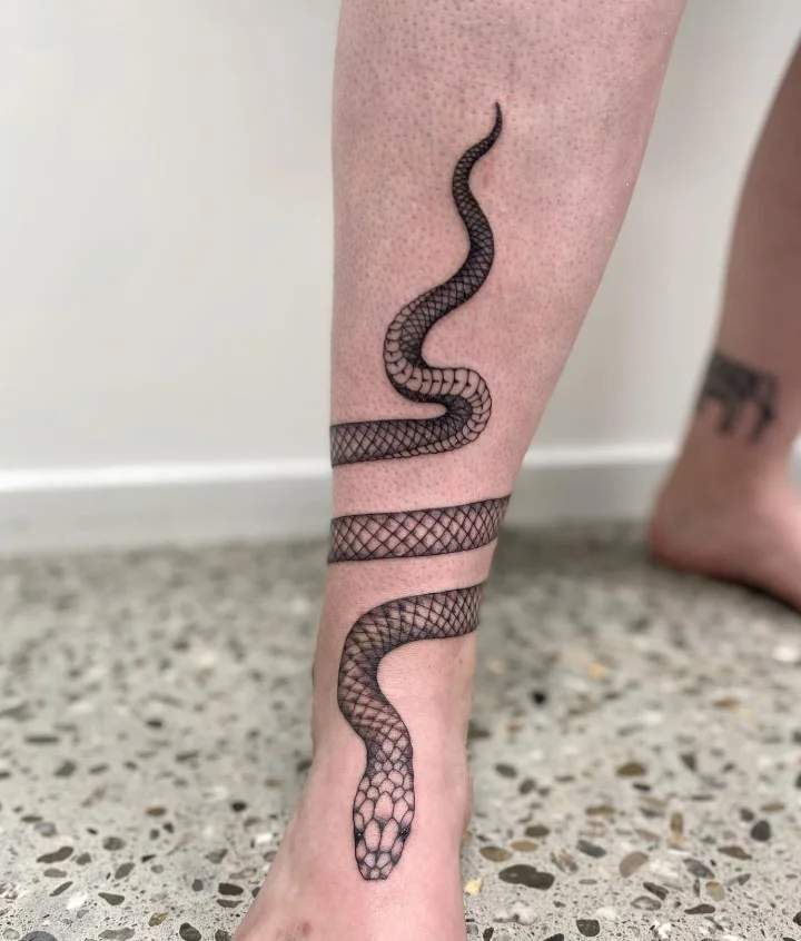Fine line snake ankle tattoo featuring a slender minimalist snake outline curving naturally around the ankle