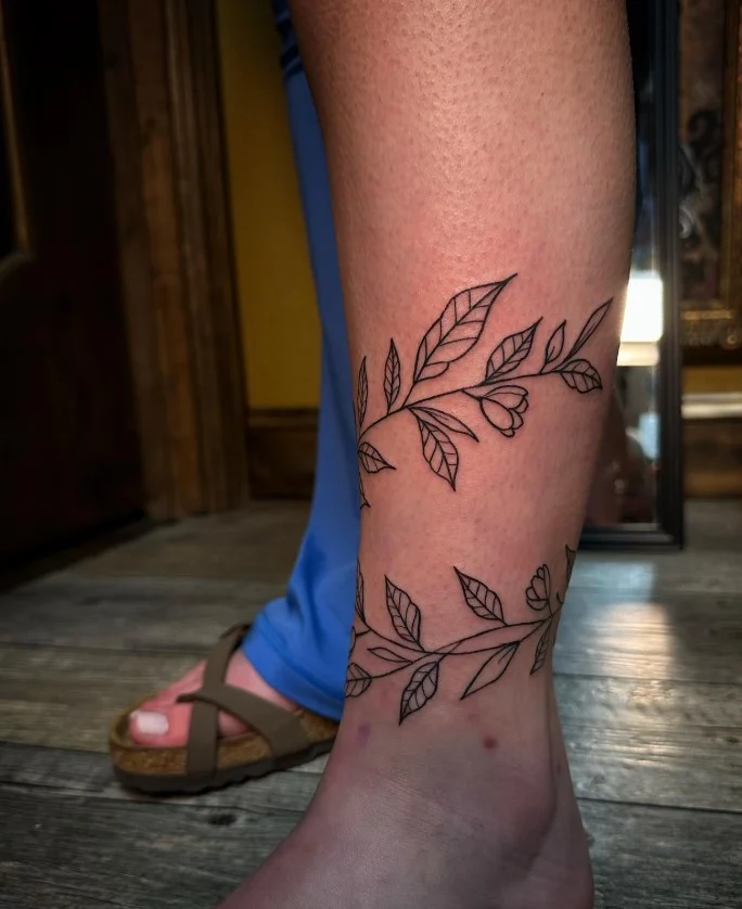 Fine line vine ankle tattoo featuring a slender curved stem with small leaves following the natural shape of the ankle