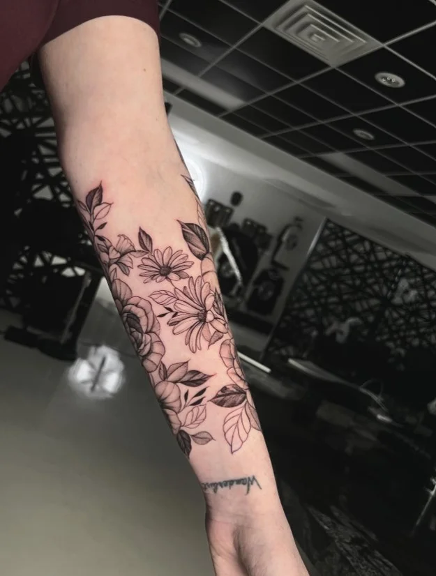 Large floral half-sleeve forearm tattoo featuring layered flowers, leaves, and flowing stems