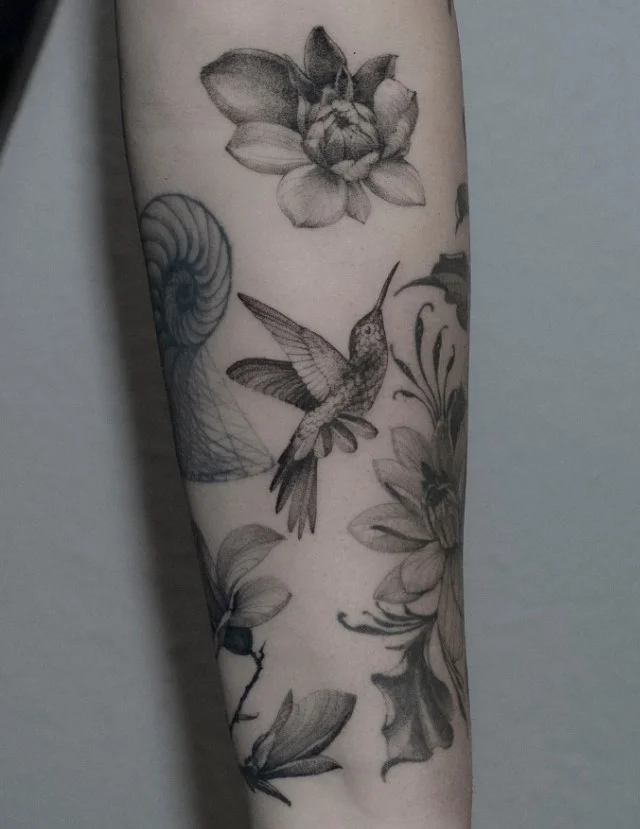 Hummingbird surrounded by delicate flowers and leafy branches wrapping around a woman's forearm