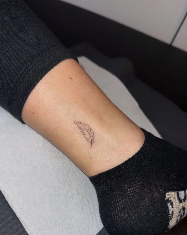 Delicate small ankle tattoo featuring a slender feather outline positioned vertically beside the ankle bone