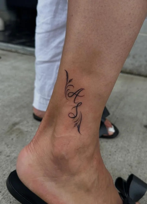 Small ankle tattoo featuring a single delicate initial in minimalist lettering on the inner ankle