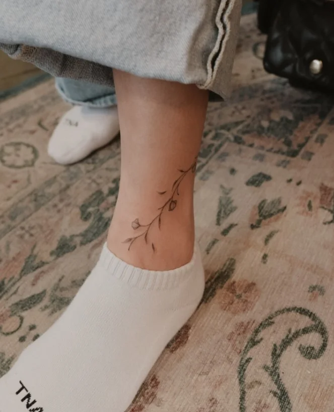 Small ankle tattoo featuring a slender curved vine with a few delicate leaves following the shape of the ankle