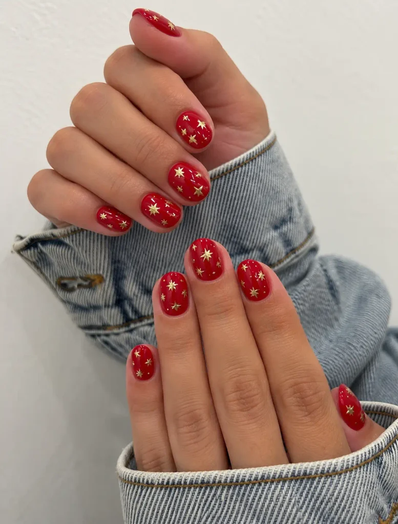 Short cherry red Christmas nails decorated with scattered gold star nail art.
