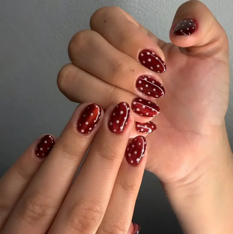 Short red Christmas nails with small white polka dots scattered across a glossy red manicure.