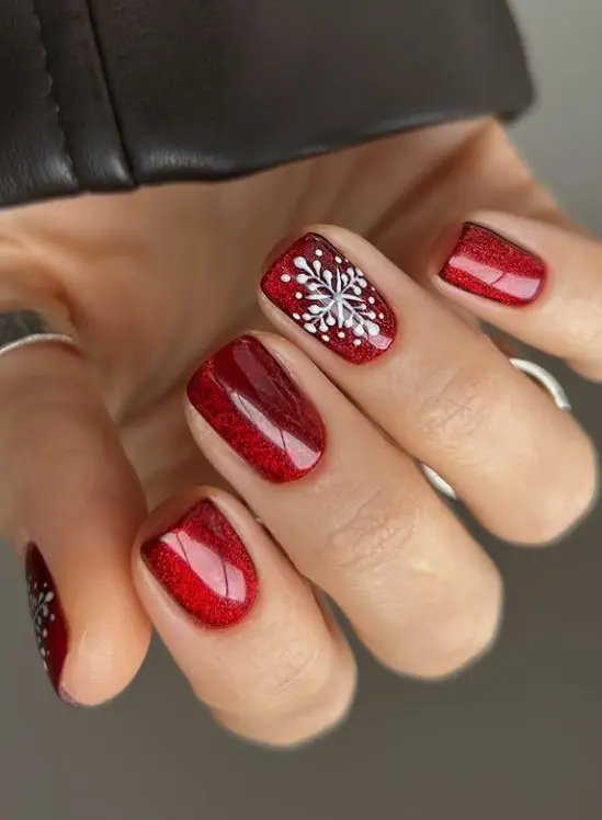 Short red Christmas nails decorated with delicate white snowflakes on an accent nail.